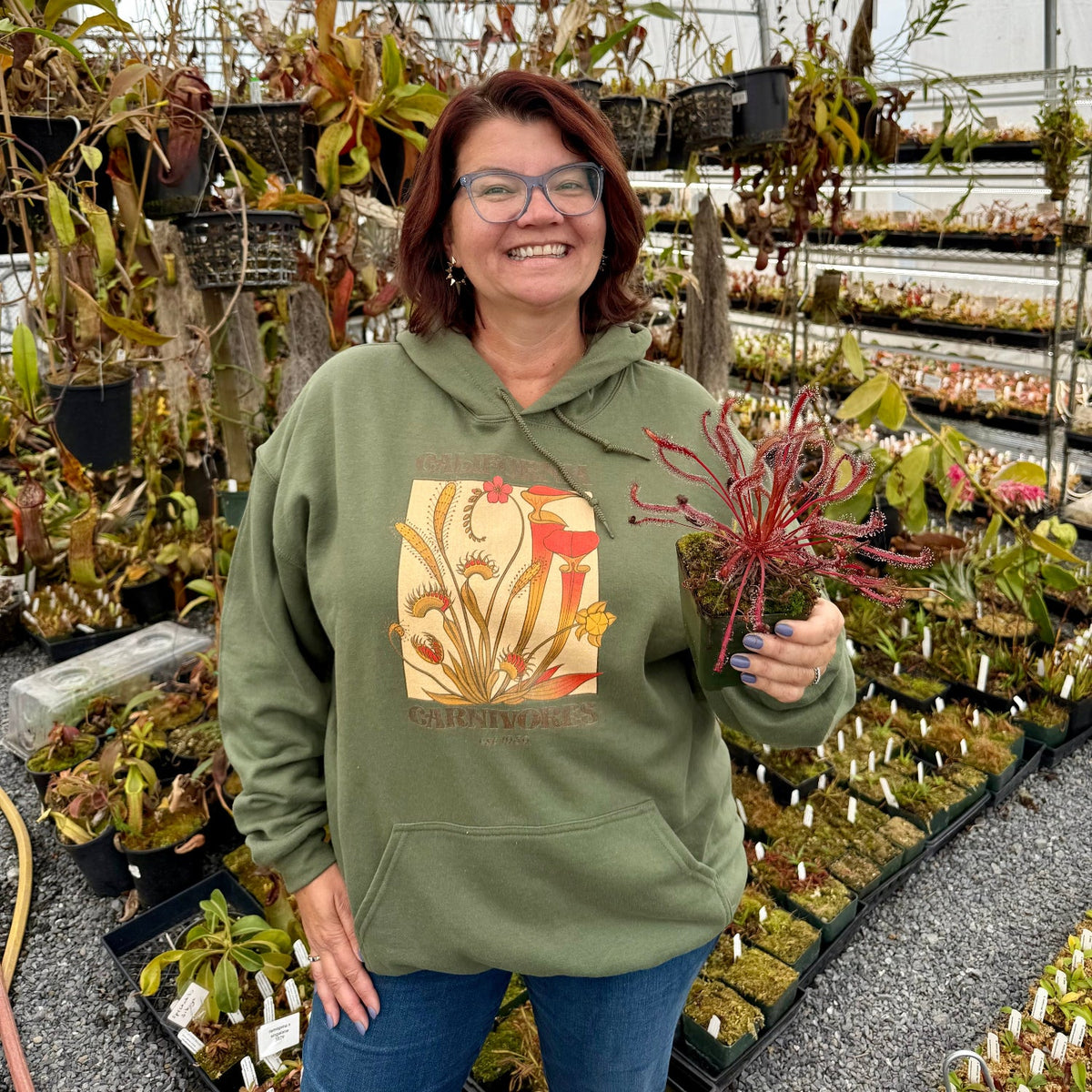 A smiling woman wearing a sage green hoodie with a colorful pitcher plant graphic stands in a greenhouse holding a red pitcher plant. Rows of potted carnivorous plants are visible in the background.