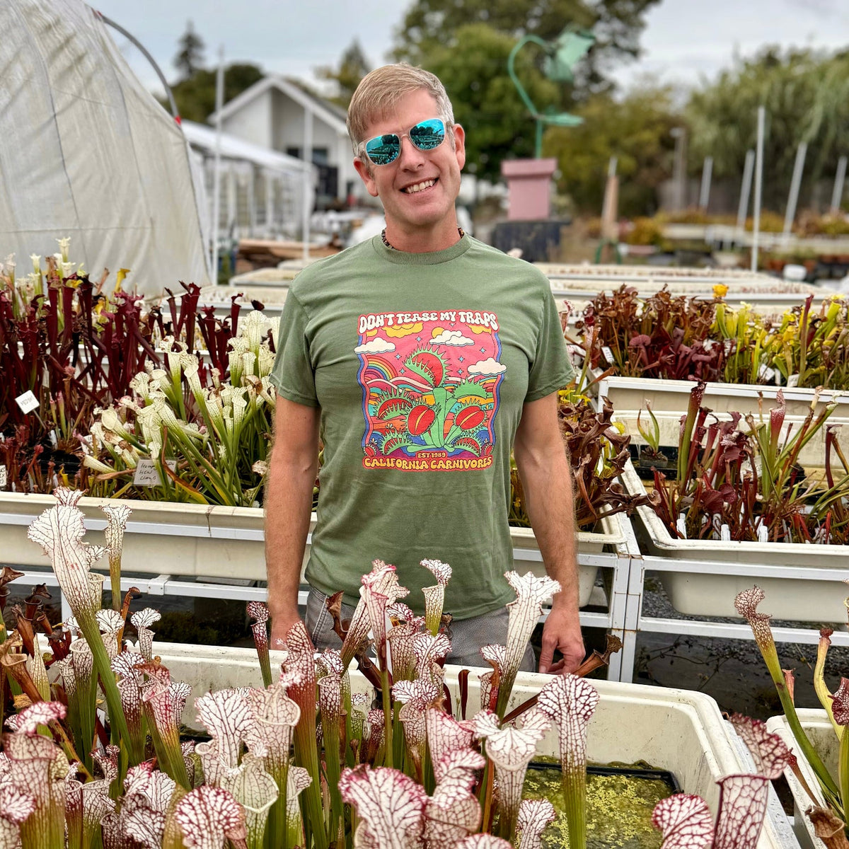 Man in sunglasses and a green shirt with "Don't tease my traps" across it in a nursery setting surrounded by Sarracenia pitchers