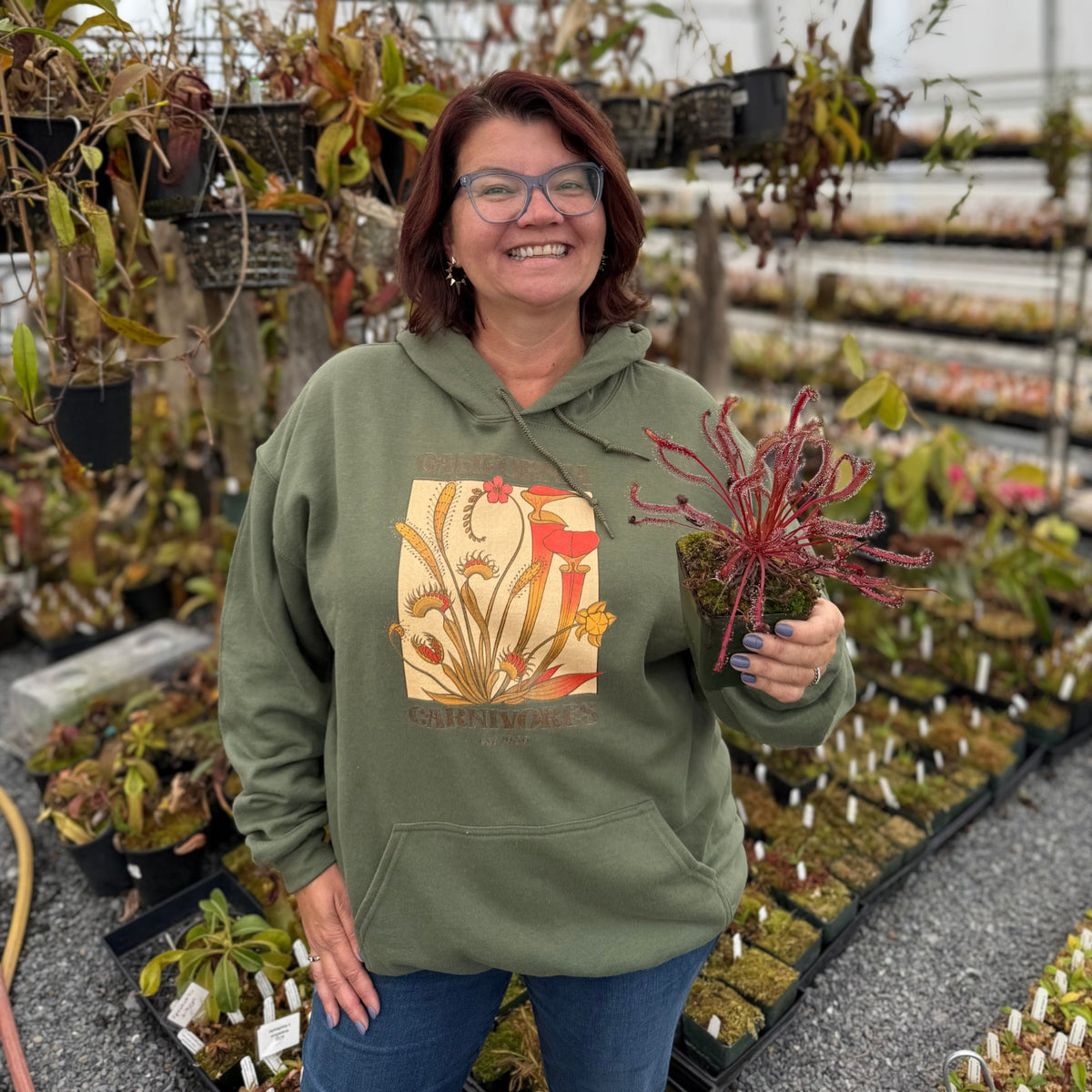 Person wearing a green hoodie with plant design, holding a plant in a greenhouse.