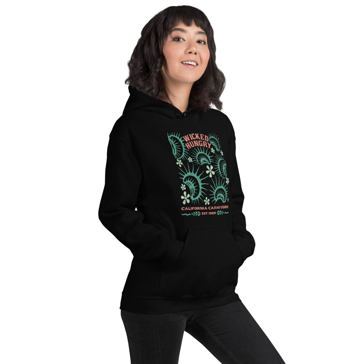 Black California Carnivores unisex hoodie with Wicked Hungry in coral, in the upper middle, surrounded by wood block print style flytraps in teal on female model