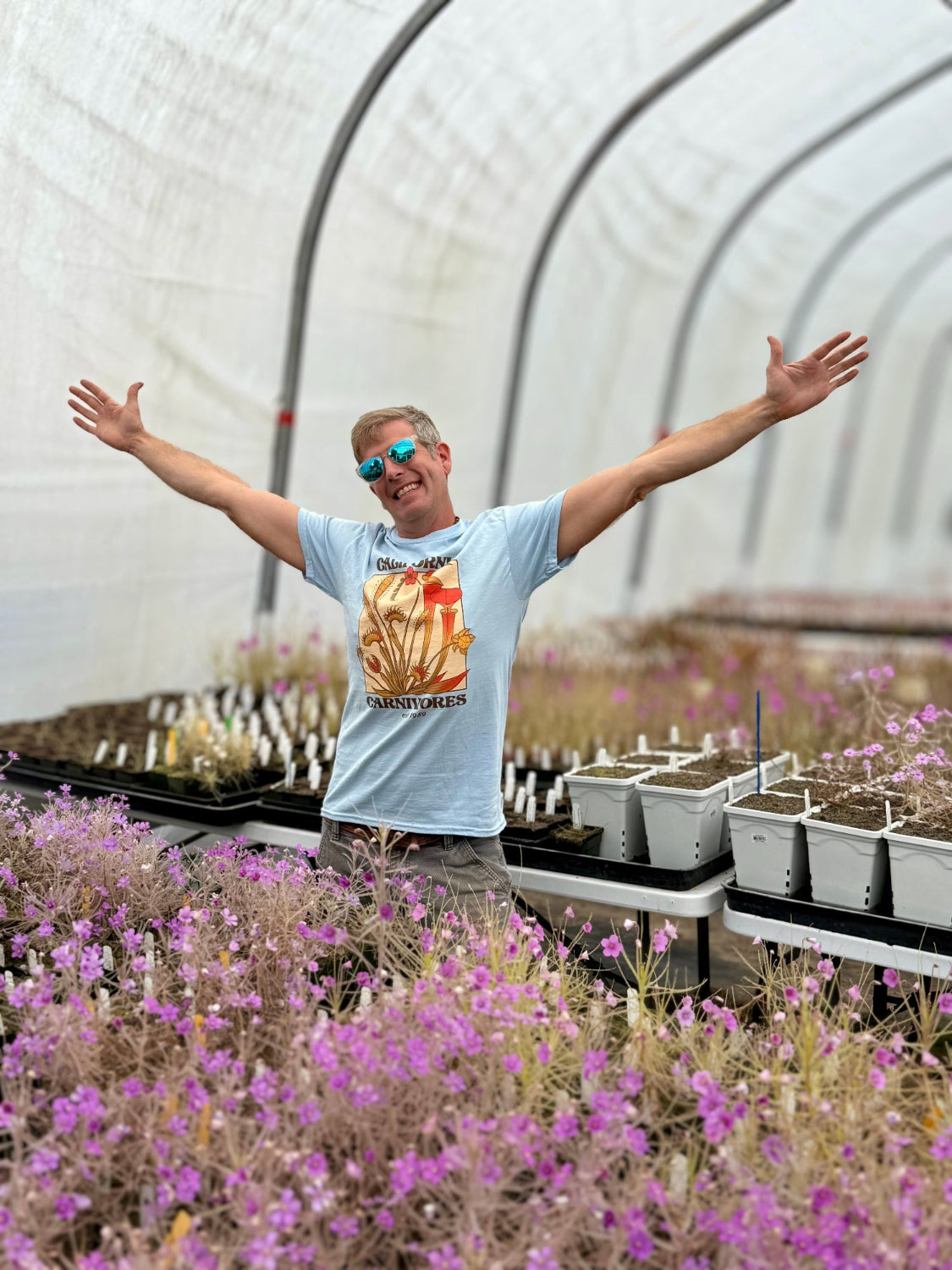 Person standing in a greenhouse with arms outstretched, surrounded by plants and seedlings.