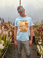 Man wearing a light blue t-shirt with a graphic design standing in a greenhouse filled with plants.